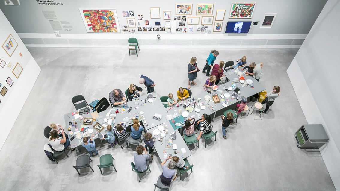 Foto: Blick von oben: Viele Kinder und Erwachsene stehen und sitzen an einem Tisch mit vielen Bastelmaterialien. Sie basteln und unterhalten sich. Viele gemalte Bilder und Zeichnungen hängen an den Wänden.
