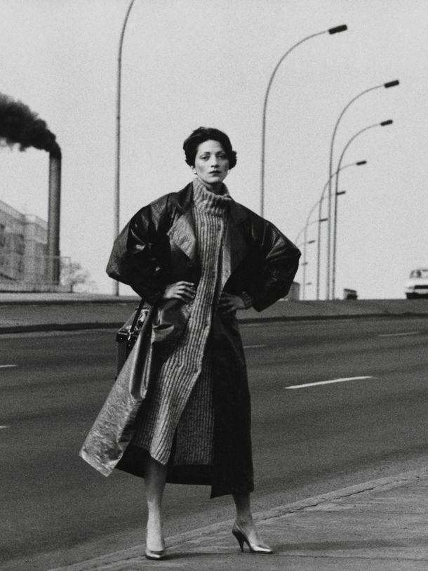 Black photograph of a woman in a coat, hands on hips, standing on a multi-lane road. In the background, streetlights and a tower from which dark smoke rises.