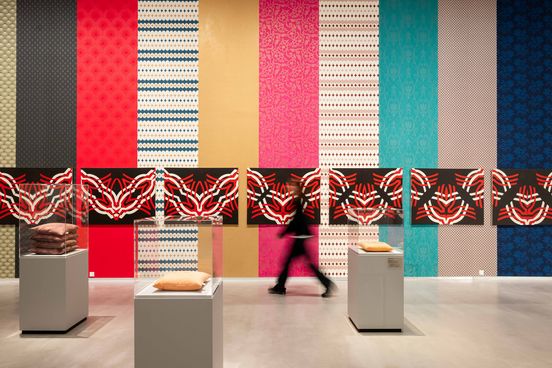 Photo: A person walks past a wall with differently patterned strips of wallpaper. In the front part of the picture there are three pedestals with glass covers, each with an embroidered cushion underneath.