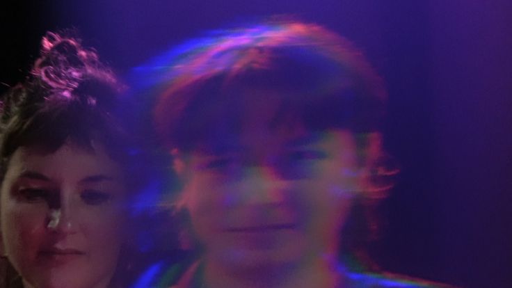 Filmstill: Das Filmstill zeigt die verschwommenen Gesichter von zwei Personen, die in lila Licht getaucht sind.