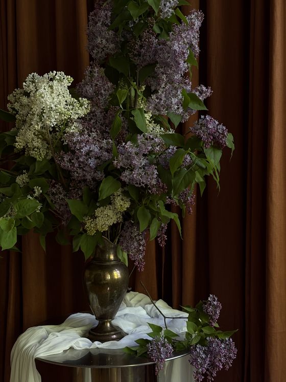 Foto: Ein üppiges Blumenbouquet steht in einer goldenen Vase vor einem bräunlichen Vorhang.