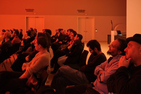Photo: Seated audience bathed in warm, reddish-orange light.