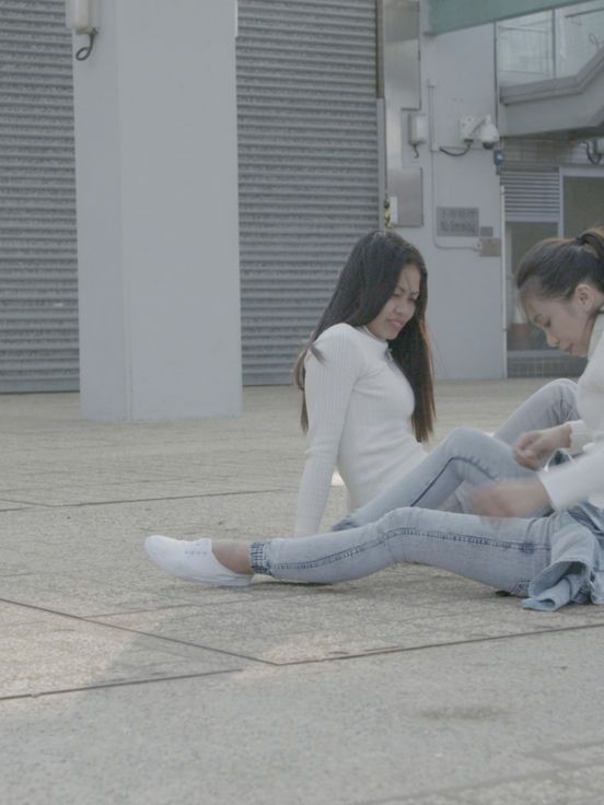 Film still: Zwei junge Frauen, die auf einem gepflasterten Außenplatz vor einem Gebäude mit grauen Rolltoren sitzen. Beide tragen helle Rollkragenpullover und Jeans. Die Frau im Vordergrund sitzt mit ausgestreckten Beinen auf dem Boden und scheint ihre Kleidung zu richten, während die andere Frau leicht hinter ihr sitzt und zu ihr hinabsieht. Die Szene ist in entsättigten, kühlen Tönen gehalten.