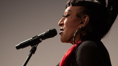 Photo: Side view of Joey Arias at the microphone, wearing a long black ponytail with a red tip.