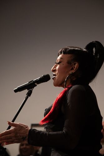 Photo: Side view of Joey Arias at the microphone, wearing a long black ponytail with a red tip.