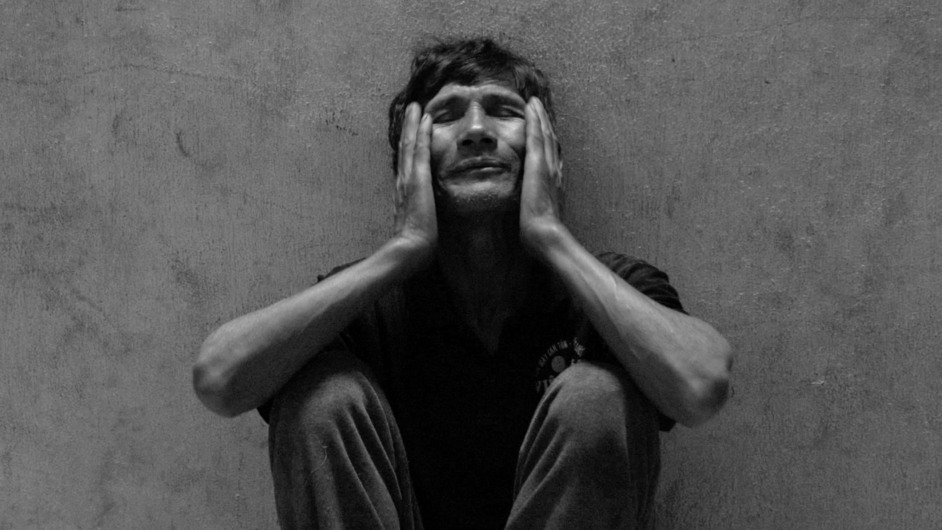 Video still: Black and white image of a person sitting on the floor leaning against a wall with both hands on their cheeks. The person's eyes are closed.