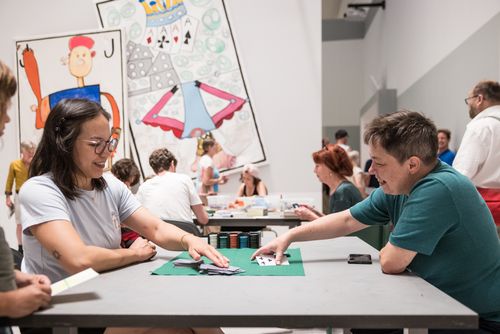 Photo: Some people are sitting at tables in the project room of the Berlinische Galerie, playing card games, doing handicrafts and chatting.