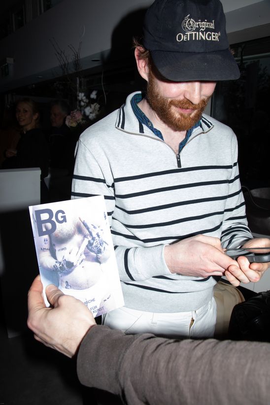 Foto: Eine Person hält die Einladungskarte zur Ausstellung in die Kamera, der Hintergrund zeigt eine weitere Person am Handy.