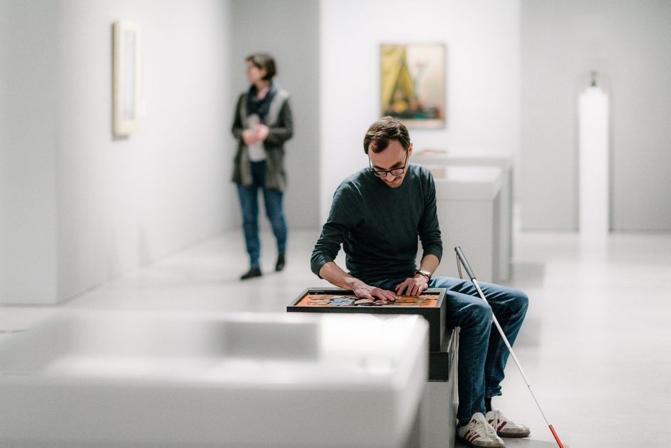 Photo: Visitor sitting on a seat with a white cane touching a tactile model in the exhibition room.
