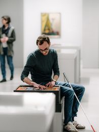 Foto: Besucher auf Sitzgelegenheit mit Blindenlangstock betastet ein Tastmodell im Ausstellungsraum.