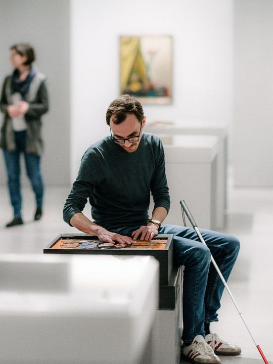 Photo: Visitor sitting on a seat with a white cane touching a tactile model in the exhibition room.