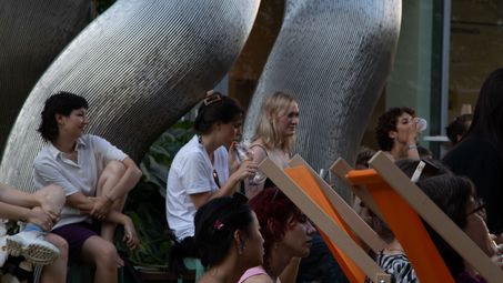 Foto: Seitlicher Blick auf einige Personen, die vor einer großen silbernen Skulptur auf dem Vorplatz der Berlinischen Galerie sitzen.