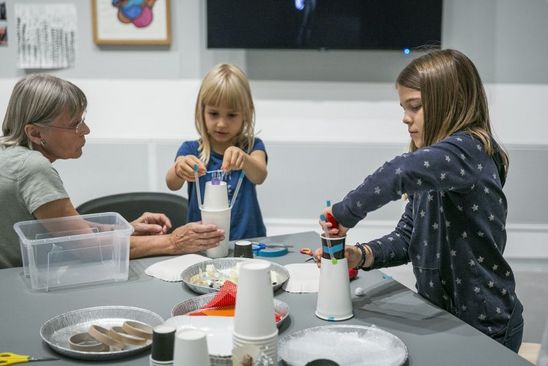 Photo: Two children and an adult are sitting around a table with handicraft materials. The children are making things out of paper cups.