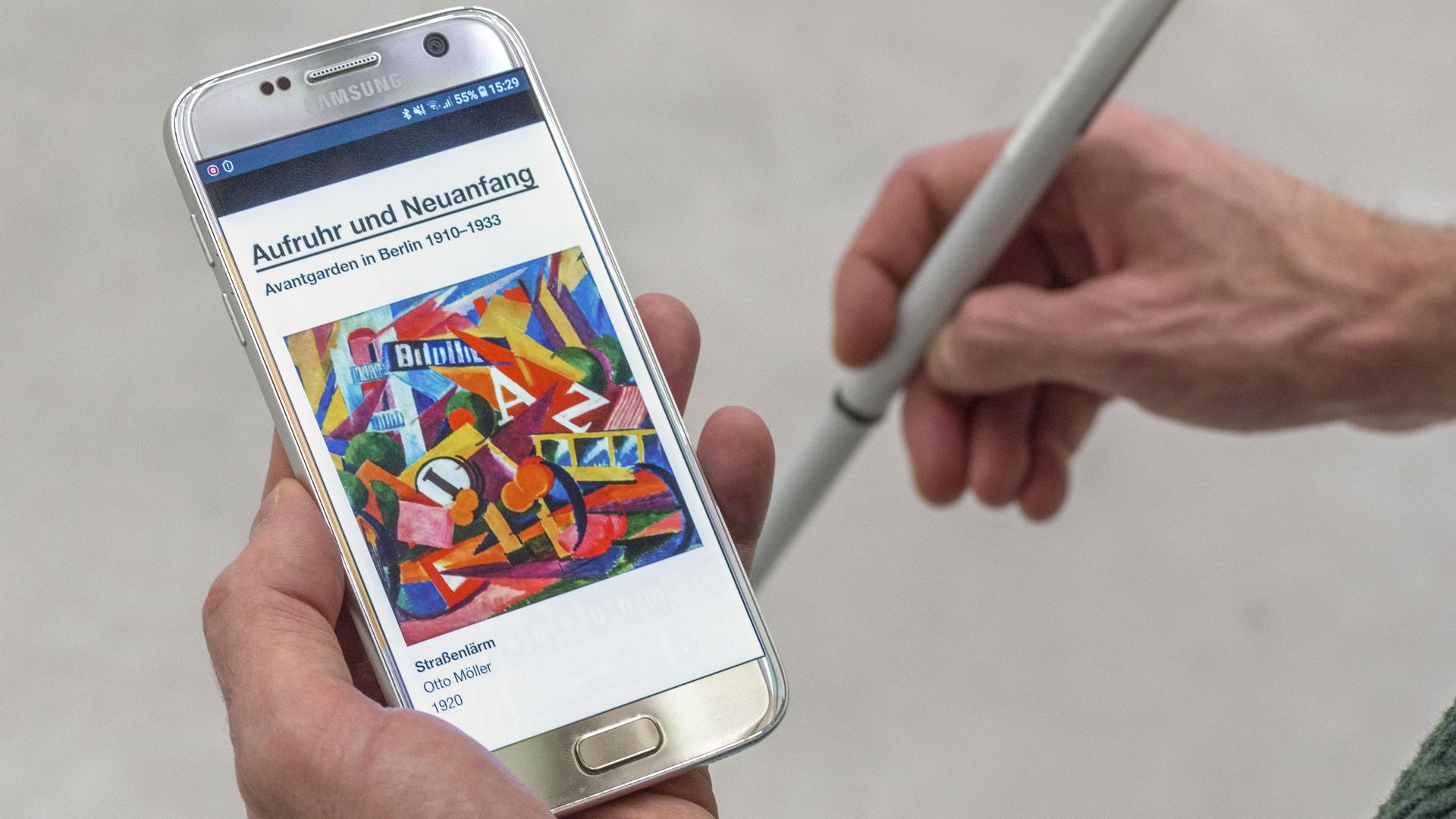 Besucher:in mit Blindenlangstock und Smartphone in der Hand. Es zeigt einen Audioguide mit der Abbildung eines Kunstwerkes. 