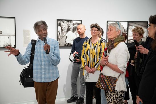 Foto: Akinbode Akinbiyi mit Mikrofon in der Hand gibt eine Tour durch die Ausstellung. Auf der rechten Bildseite sind einige Teilnehmende zu sehen.