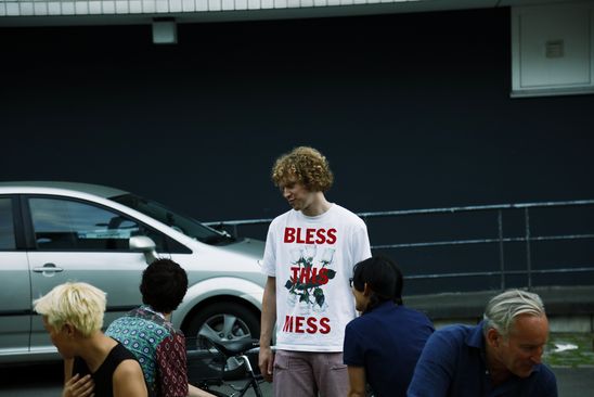 Eine Person steht draußen vor dem Museum und unterhält sich, während um sie herum weitere Menschen sitzen. Ihr weißes Tshirt trägt in rot die Aufschrift: „Bless This Mess“.