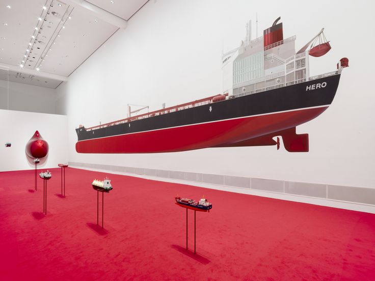 Foto: Blick in den Ausstellungsraum mit einem großen an die Wand gemalten Öltanker und weiteren Installationen von Monira Al Qadiri. Im gesamten Ausstellungsraum liegt ein leuchtend roter Teppich.