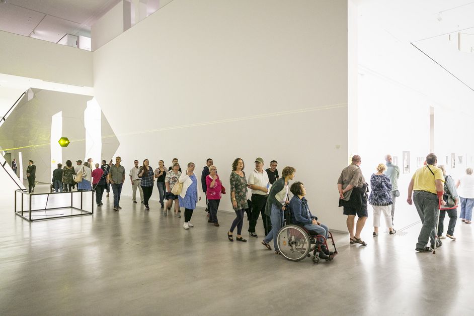 Foto: Viele Menschen unterschiedlichen Alters, mit und ohne Gehbehinderungen laufen durch einen Ausstellungsraum mit einer Laserinstallation.