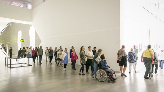 Foto: Viele Menschen unterschiedlichen Alters, mit und ohne Gehbehinderungen laufen durch einen Ausstellungsraum mit einer Laserinstallation.