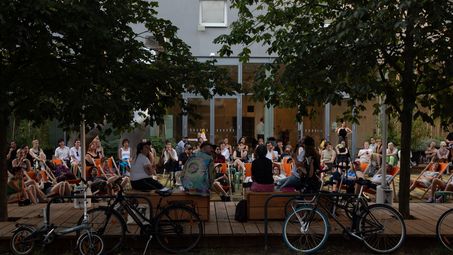 Foto: Viele Menschen sitzen auf bunten Stühlen, Hockern oder auf dem Boden auf dem Vorplatz der Berlinischen Galerie und lauschen einem Podium aus fünf Personen.