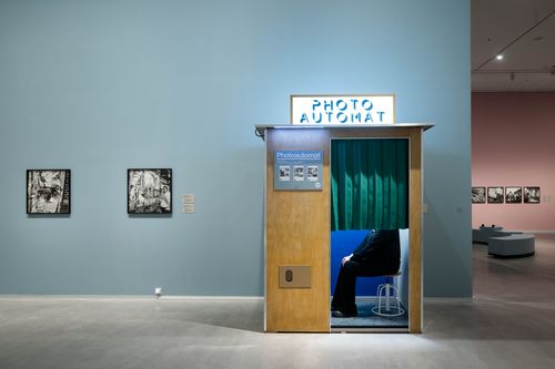 Exhibition view "Akinbode Akinbiyi": View of a photo machine in which a person is sitting behind a dark green curtain. To the left of the machine hang two black and white photographs. To the right of the machine is another exhibition space with seating and more photographs.