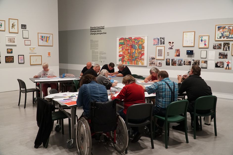 Foto: Menschen sitzen und arbeiten an Tischen mit künstlerischen Materialien. An den Wänden hängen viele kleine Kunstwerke.