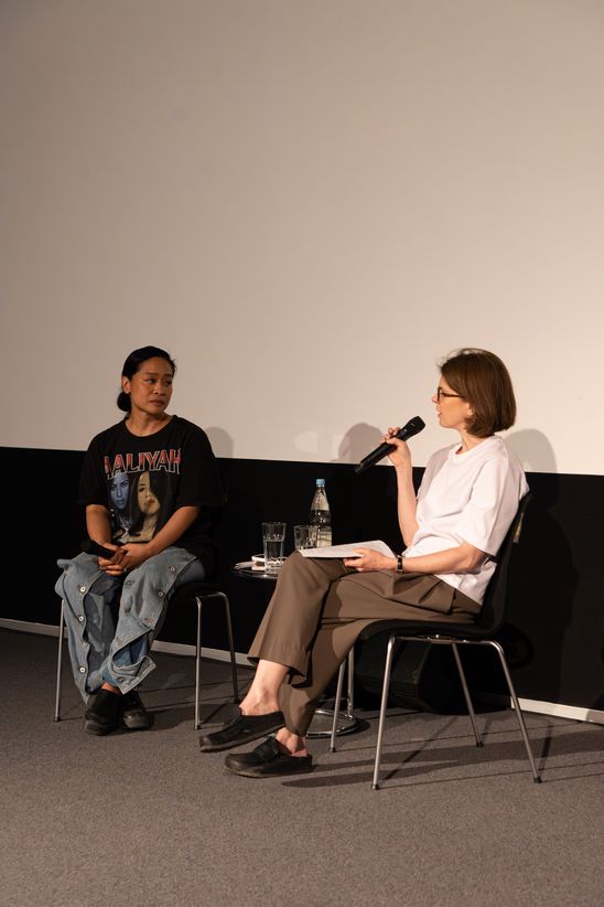 Foto: Stephanie Comilang und Anne Bitterwolf sitzen auf Stühlen vor einer weißen Leinwand, Anne Bitterwolf hält ein Mikrofon in der Hand.