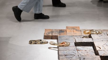 Photo: The lower part of the photo shows bricks lying next to each other with imprints embedded in them. Next to and on top of the bricks is an imprint of a foot made of semi-precious stones and synthetic resin. The legs of two people walking past can be seen in the background.