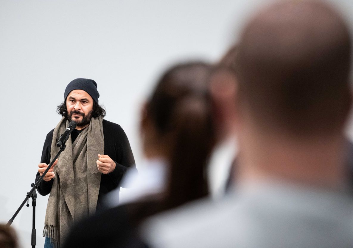 Foto: Der Künstler Kader Attia steht vor einem Mikrofon und spricht. Er trägt dunkle Kleidung, einen sandfarbenen Schal und eine dunkle Mütze. Im rechten Bildvordergrund sind verschwommen zwei Personen von hinten zu sehen.
