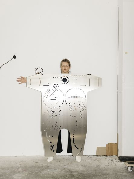 Foto: Das Porträt zeigt die Künstlerin Mariechen Danz hinter einer ihrer großen, silbernen Skulpturen. Diese ähnelt einem Torso und besteht aus Aluminium, in das kleine geometrische Formen eingestanzt sind. Mariechen Danz wird von der Skulptur fast ganz verdeckt, nur der Kopf, eine Hand, und die Füße ragen hinaus.