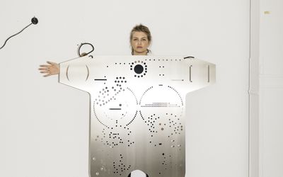 Foto: Das Porträt zeigt die Künstlerin Mariechen Danz hinter einer ihrer großen, silbernen Skulpturen. Diese ähnelt einem Torso und besteht aus Aluminium, in das kleine geometrische Formen eingestanzt sind. Mariechen Danz wird von der Skulptur fast ganz verdeckt, nur der Kopf, eine Hand, und die Füße ragen hinaus.
