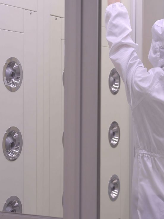 Filmstill: Rückenansicht einer Person in weißem Schutz-Overall, die in einem abgeschlossenen Raum steht, an dessen langer Wand sich chromfarbene Wasserdrüsen befinden.