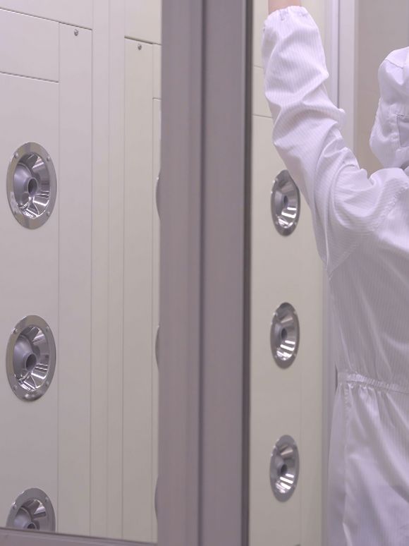 Filmstill: Rückenansicht einer Person in weißem Schutz-Overall, die in einem abgeschlossenen Raum steht, an dessen langer Wand sich chromfarbene Wasserdrüsen befinden.