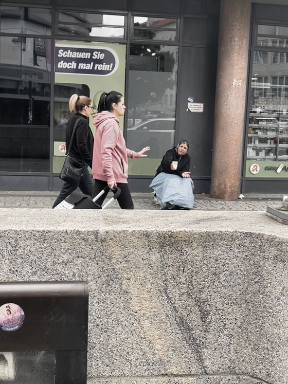 Eine Frau sitzt bettelnd auf dem Boden. An ihr vorbei laufen zwei weitere Frauen. Eine der beiden macht eine abwehrende Geste mit der Hand.