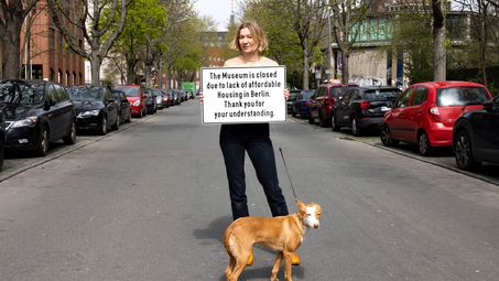 Cem A., "Thank you for your understanding", 2023 A person stands with a dog on a Berlin street holding a sign that reads “The Museum is closed due to lack of affordable Housing in Berlin. Thank you for your understanding.” Parked cars, building facades, and trees are visible in the background.