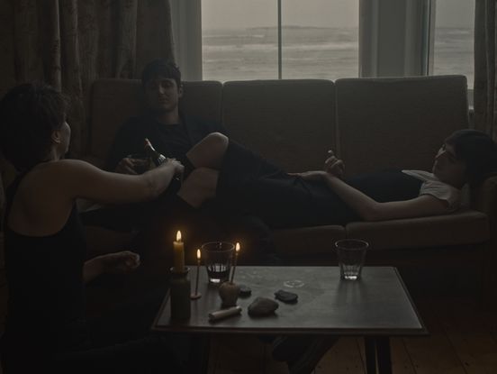 Film-Still: Blick in einen Raum mit Fenster zum Meer. Auf einem Sofa am Fenster liegt eine Person und hat die Beine auf den Oberschenkeln einer anderen Person abgelegt. Gegenüber sitzt eine dritte Person und schenkt ein Glas ein.
