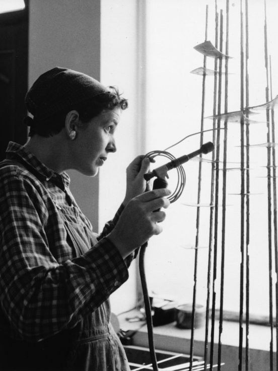 Black and white photo: Side view of Brigitte Meier-Denninghoff working on one of her sculptures in front of a window with a soldering iron in her hand.