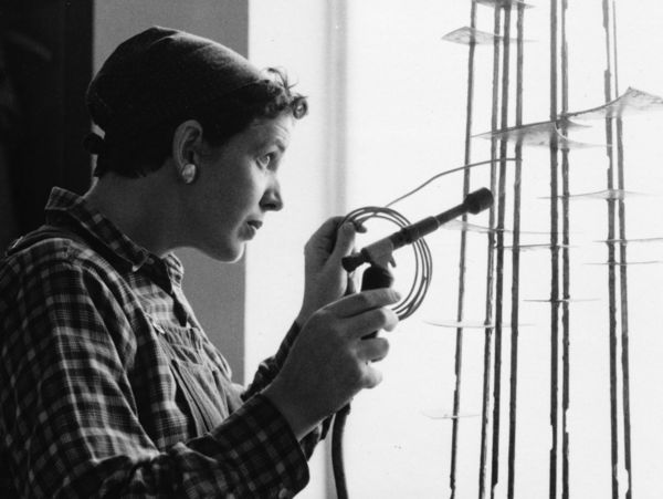 Brigitte Meier-Denninghoff, München 1957 Schwarz-Weiß-Foto: Seitliche Aufnahme von Brigitte Meier-Denninghoff, die vor einem Fenster mit Lötkolben in der Hand an einer ihrer Skulpturen arbeitet.