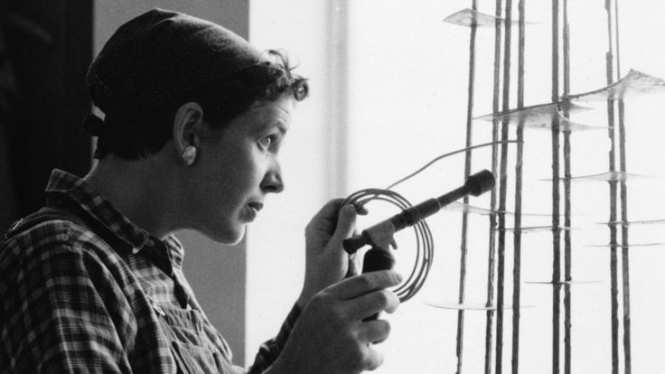 Schwarz-Weiß-Foto: Seitliche Aufnahme von Brigitte Meier-Denninghoff, die vor einem Fenster mit Lötkolben in der Hand an einer ihrer Skulpturen arbeitet.