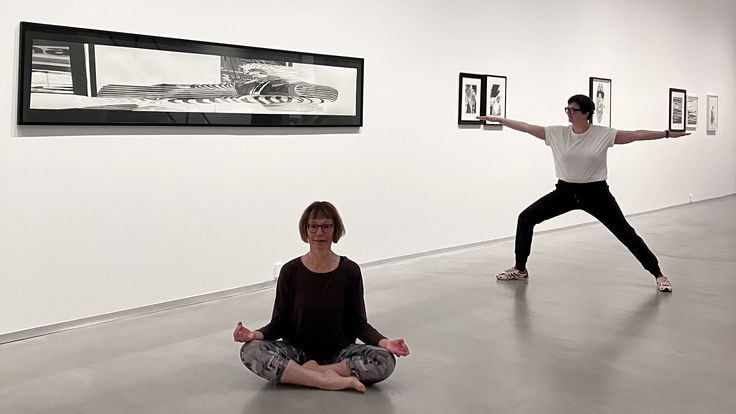 Foto: Zwei Personen, teilweise in Yoga-Kleidung, stehen in Yoga-Posen vor einer Wand mit gerahmten Zeichnungen von Marc Brandenburg.