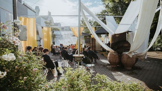 Besucher*innen sitzen auf Stühlen auf dem Parkplatz und blicken auf eine Bühne