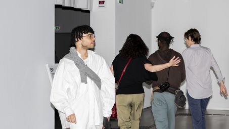 Photo with flash: Front view of a person in white clothing and glittering silver shoes walking out of an exhibition room, while three people walk into the room in the background.