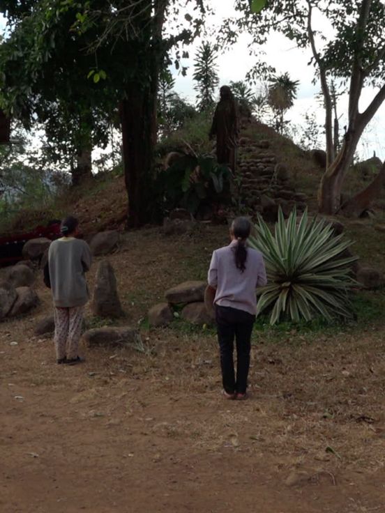 Video still: Back view of three people with their hair tied together, standing in front of a small hill with stone steps.