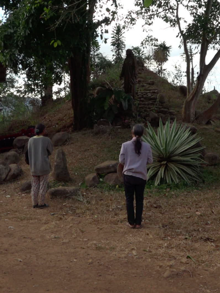 Video-Still: Rückenansicht von drei Personen mit zusammengebundenen Haaren, die vor einem kleinen Hügel mit steinernen Stufen stehen.
