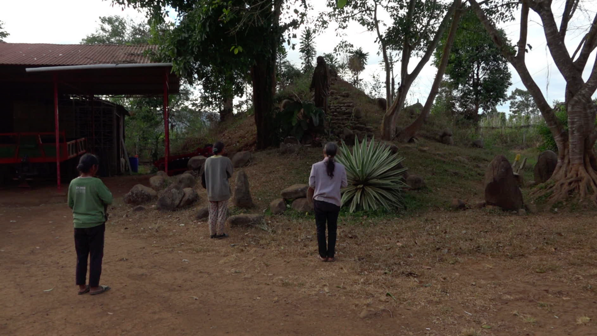 Video still: Back view of three people with their hair tied together, standing in front of a small hill with stone steps.