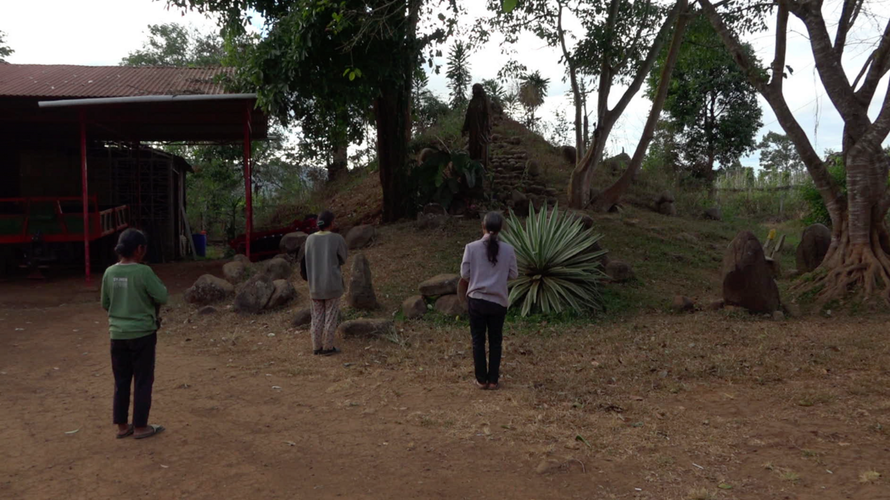 Video-Still: Rückenansicht von drei Personen mit zusammengebundenen Haaren, die vor einem kleinen Hügel mit steinernen Stufen stehen.