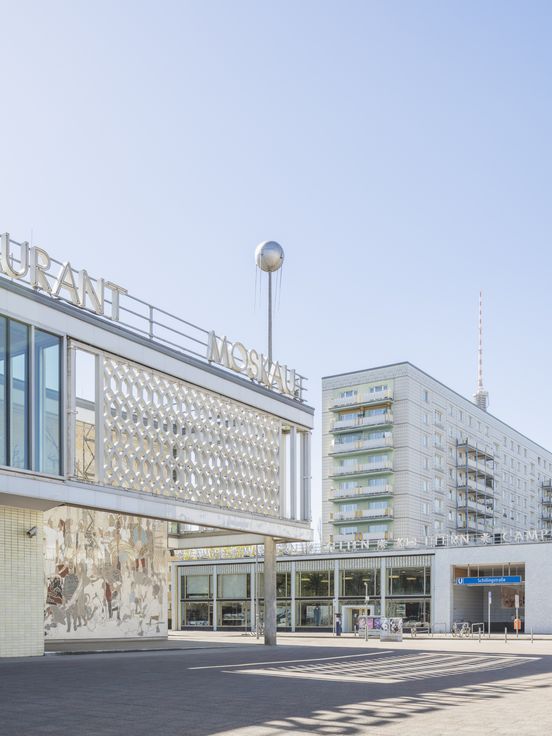 Ansicht des Café Moskau in Berlin mit dem Schriftzug „MOSKAU“, ornamentaler Fassade und umliegender DDR-Architektur unter blauem Himmel