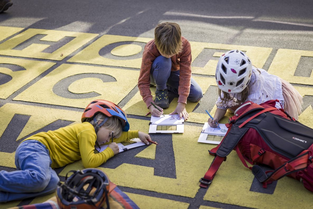 Abdrücke und Eindrücke der Stadt Drei Kinder untersuchen ein Bodenkunstwerk, auf dem sie sitzen: Graue Buchstaben auf gelbem Untergrund ergeben Namen von Künstler:innen.