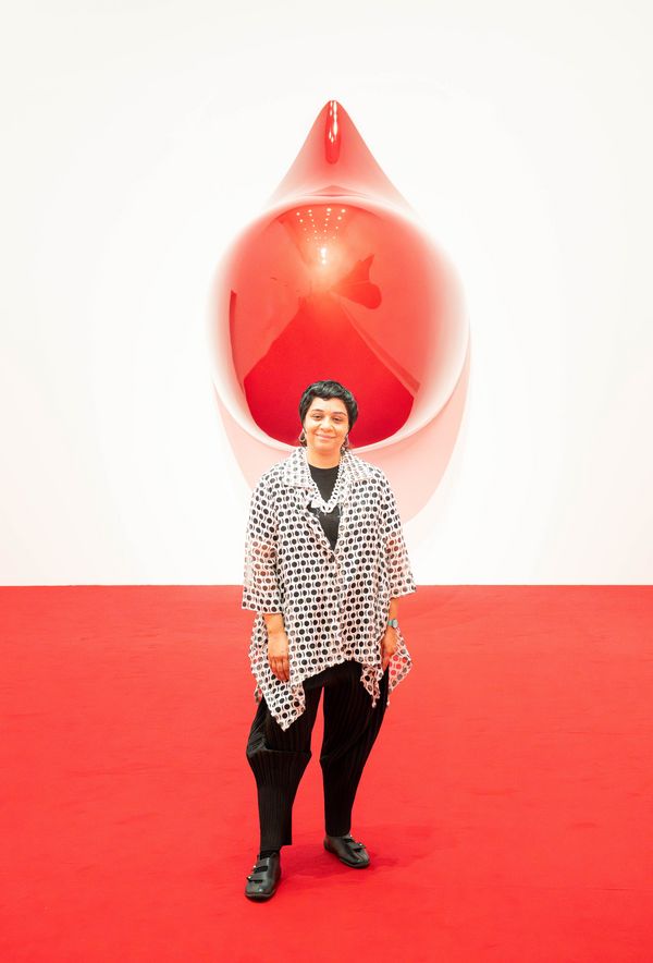 Porträt der Künstlerin Monira Al Qadiri in ihrer Ausstellung. Sie steht auf einem roten Teppich vor einer großen, glänzenden, roten Skulptur an der Wand.