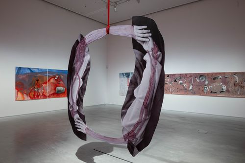 Exhibition view "Özlem Altın": An oval installation of four touching arms hangs on a red rope in the exhibition space. Three paintings can be seen in the background. 